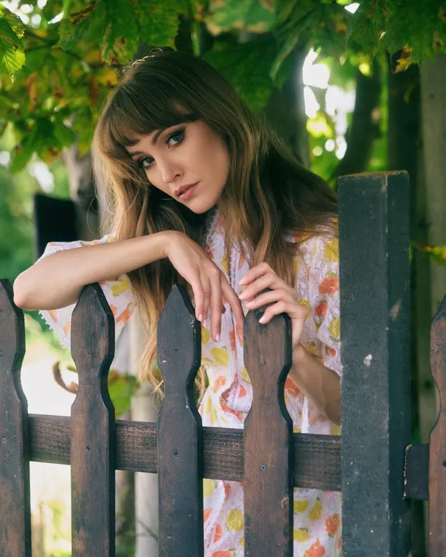Wiktoria in floral blouse leaning on a wooden fence among leaves, portrait session Rzeszów